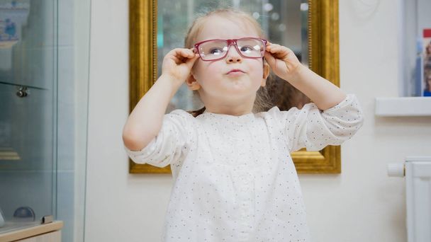 Menina tenta novos óculos perto do espelho - compras na clínica oftalmológica
 - Foto, Imagem