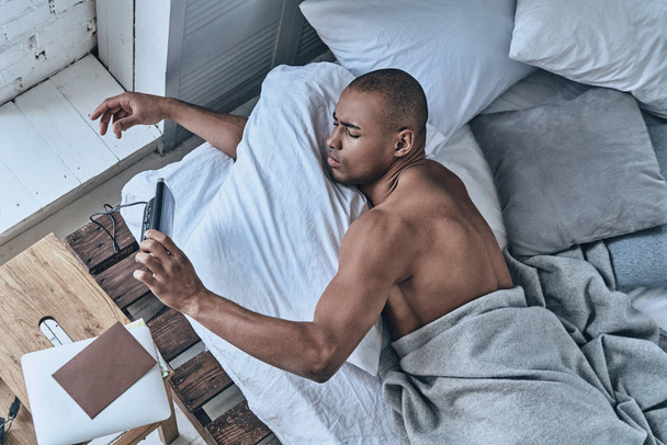 young African man turning off alarm clock while lying in bed at home  - Foto, Bild
