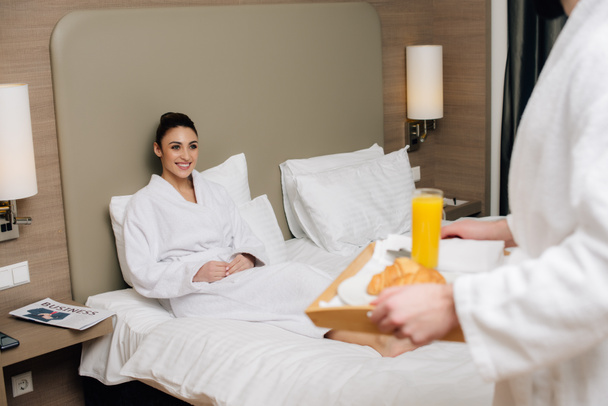 recortado disparo de hombre llevando bandeja con sabroso desayuno para la novia mientras ella sentado en la cama en la suite del hotel
 - Foto, Imagen