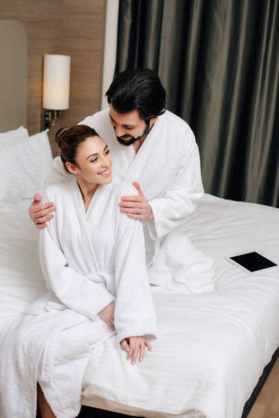 όμορφο ζευγάρι στην αγκαλιά στο κρεβάτι σε σουίτα ξενοδοχείου μπουρνούζια - Φωτογραφία, εικόνα