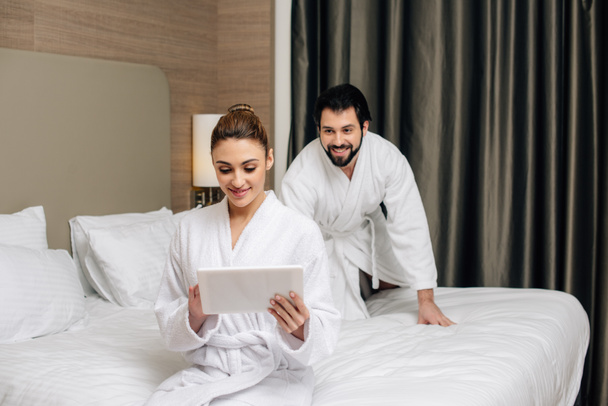 bela mulher em roupão de banho usando tablet na cama na suíte do hotel, enquanto seu namorado brincalhão sorrateiramente para ela por trás
 - Foto, Imagem