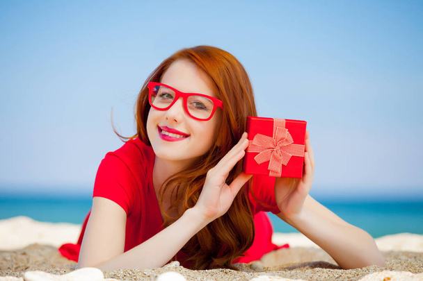 Young redhead girl in red dress with gift box have a rest on summertime sea beach - Foto, imagen