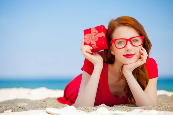 Jeune fille rousse en robe rouge avec boîte cadeau se reposer sur la plage de la mer d'été
 - Photo, image