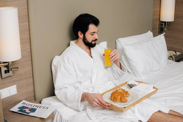 schöner Mann im Bademantel mit Croissants und Saft auf Tablett auf dem Bett in der Hotelsuite - Foto, Bild