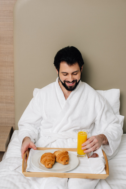 ホテルのスイート ルームのベッドの上に座ってトレイの上に食べ物をバスローブで幸せな男 - 写真・画像