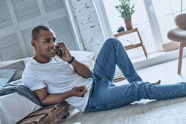 Handsome young African man talking on phone while sitting on floor at home  - Φωτογραφία, εικόνα