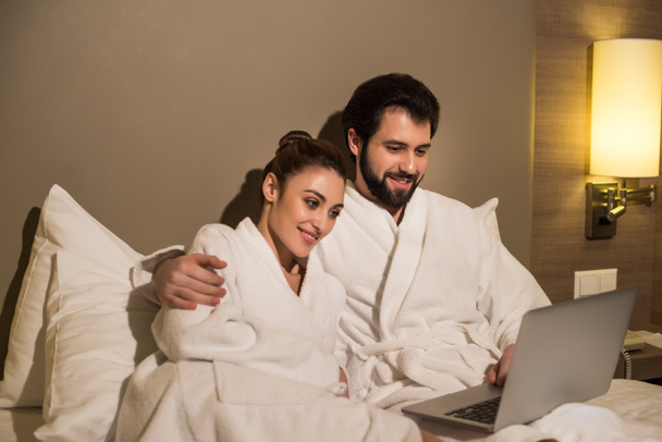 belo casal em roupões de banho usando laptop juntos na cama da suíte do hotel
 - Foto, Imagem