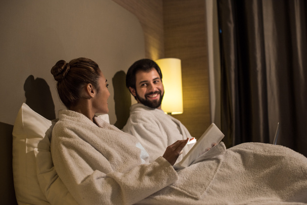 ευτυχισμένο ζευγάρι σε μπουρνούζια χρησιμοποιώντας συσκευές στο κρεβάτι του ξενοδοχείου suite - Φωτογραφία, εικόνα