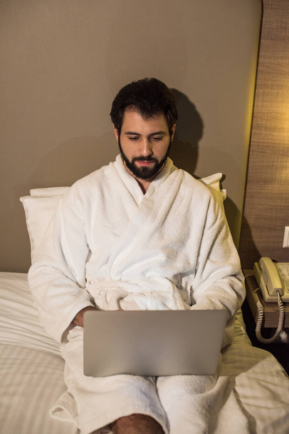 hombre guapo en albornoz usando el ordenador portátil en la cama en la suite del hotel por la noche
 - Foto, Imagen