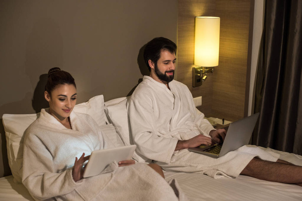 beautiful couple in bathrobes using devices in bed of hotel suite - Photo, Image