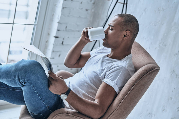 Handsome young African man reading newspaper and drinking coffee while sitting in armchair at home  - Photo, Image