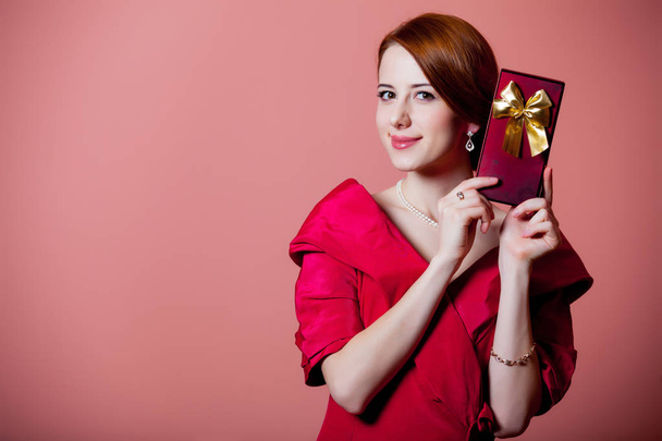Young redhead woman in red Victorian epoch clothes with gift box on pink background - Photo, Image