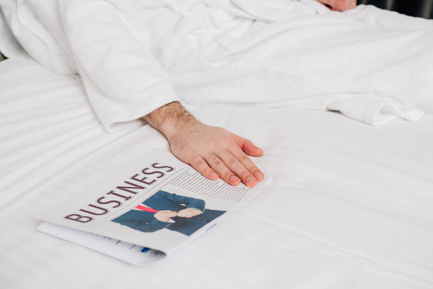 ビジネス新聞でベッドの上に横たわるバスローブの男のショットをトリミング - 写真・画像