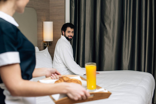 обрезанный снимок горничной в форме, держащей поднос с круассанами и соком для гостя отеля, пока он сидит на кровати
 - Фото, изображение