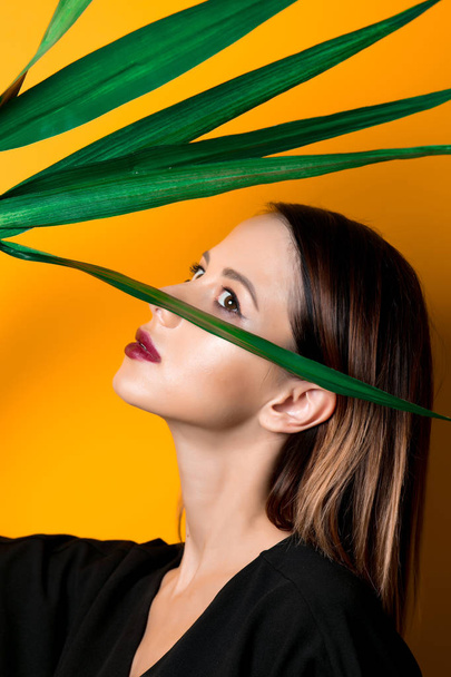 Beautiful woman in dark clothes with palm leaf on yellow color background - Foto, immagini