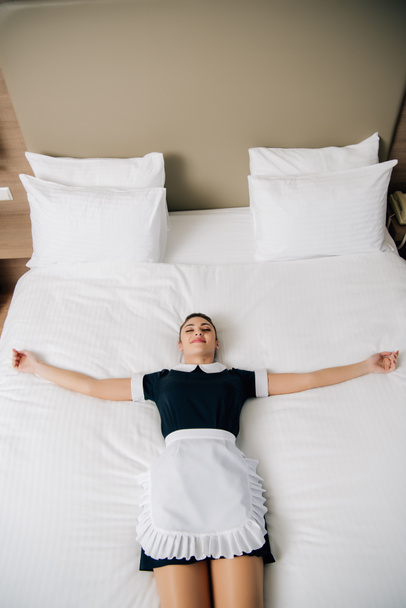 high angle view of young attractive maid in uniform lying on bed at hotel suite - Fotó, kép