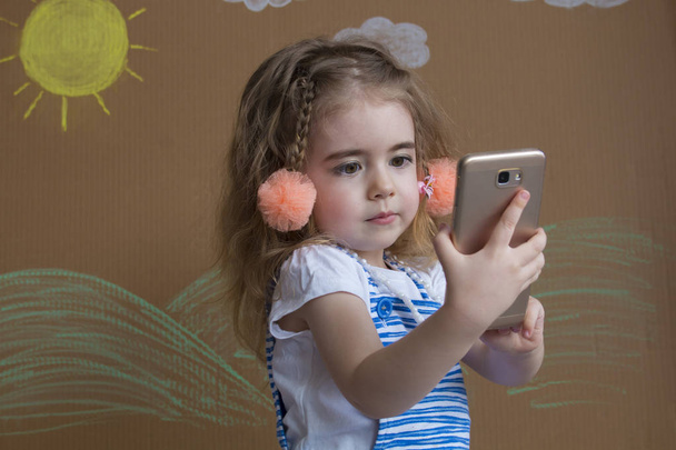Emocionální portrét drobná světlovlasá dívka a copánky, držící chytrý telefon v ruce. Pozadí malovaný slunce a bílé mraky - Fotografie, Obrázek