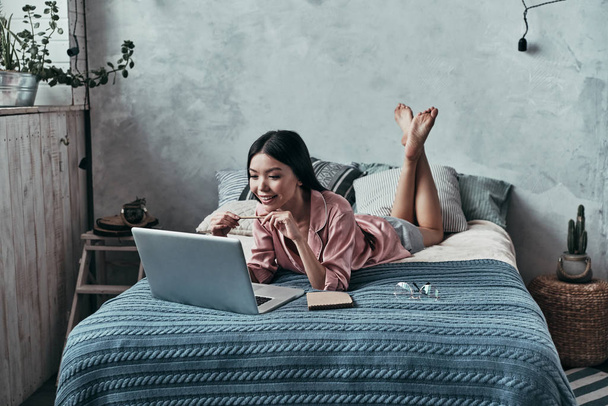 Lazy young woman using computer and smiling while lying on bed at home - 写真・画像