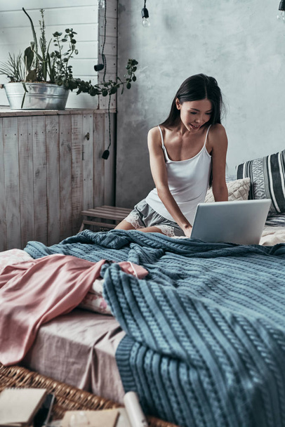 freelancer woman using computer and smiling while sitting on bed at home - Fotó, kép