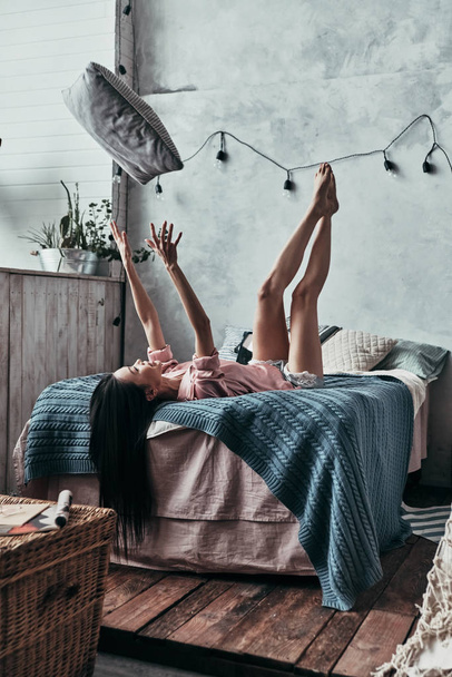 brunette young woman throwing up pillow and smiling while lying on bed at home - Fotografie, Obrázek