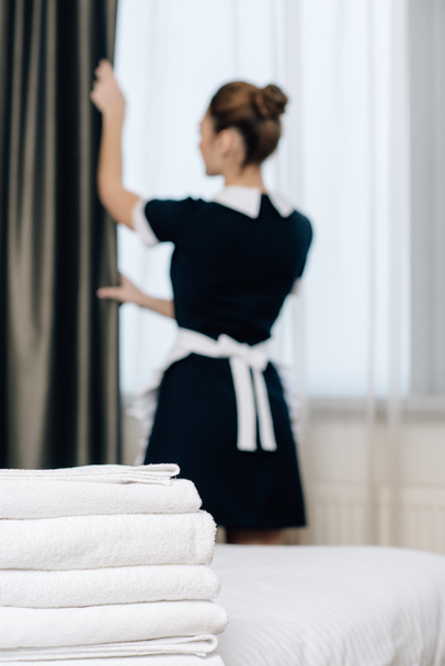 young maid in uniform shutting curtain with stack of clean towels standing on foreground - Foto, afbeelding