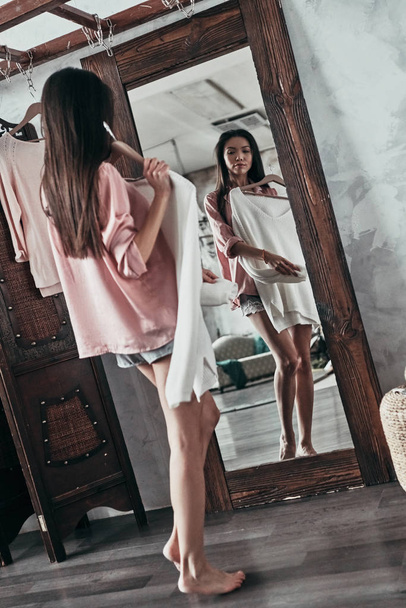 full length of attractive young woman trying on white blouse while standing in front of the mirror at home - Foto, imagen