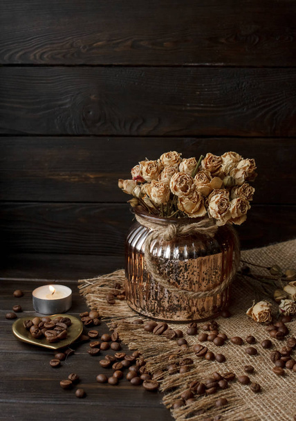 vaso de vidro de bronze com flor prensada em uma toalha de mesa. disco em forma de lareira dourada com grãos de café torrados. Arábica pura em uma mesa de madeira. Vela acesa no fundo. Café da manhã aromático
 - Foto, Imagem