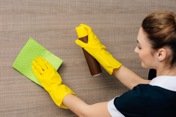 close-up shot of young maid in uniform wiping wood wall with rag and aerosol furniture cleaner - Photo, Image