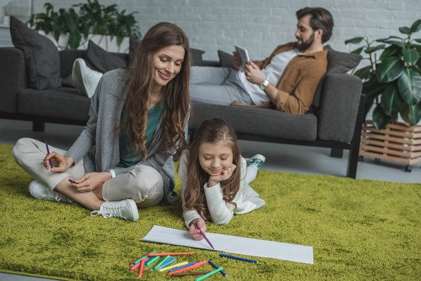 mother and daughter drawing on floor and father using tablet on sofa  - Photo, Image