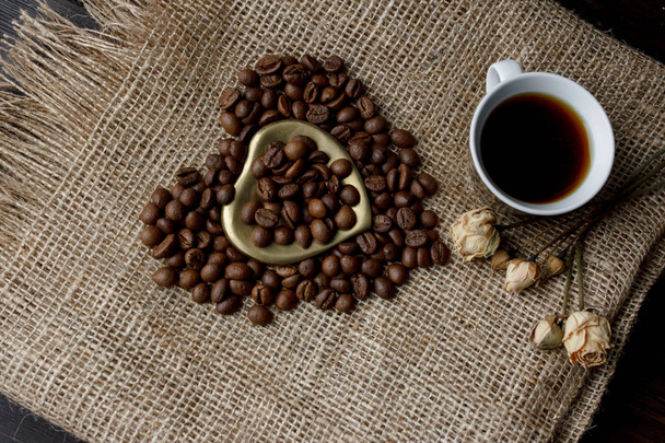 Flat lay of roasted coffee beans on a tablecloth with a golden heart shaped saucer and coffee mug. Cup of morning espresso and pressed roses. Romantic ideas - Photo, Image
