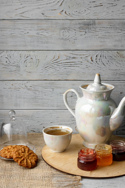 Три маленькі банки домашнього ягідного варення на дерев'яному підносі. Здорові харчові ідеї. Чайний чайник і чай з зеленого чаю на фоні. Пресовані троянди на дерев'яному столі поруч зі срібною ложкою
 - Фото, зображення