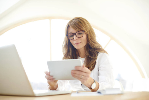 Retrato de empresária casual de meia-idade usando tablet digital enquanto se senta na mesa de escritório na frente do laptop e trabalha em novo projeto. Brainstorming no escritório
.  - Foto, Imagem