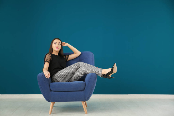 Beautiful young woman sitting in comfortable armchair against color wall - Fotografie, Obrázek