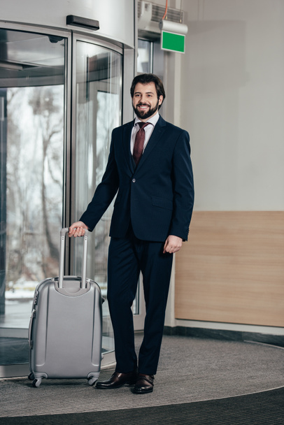 smiling handsome businessman with luggage going out of hotel - Foto, afbeelding