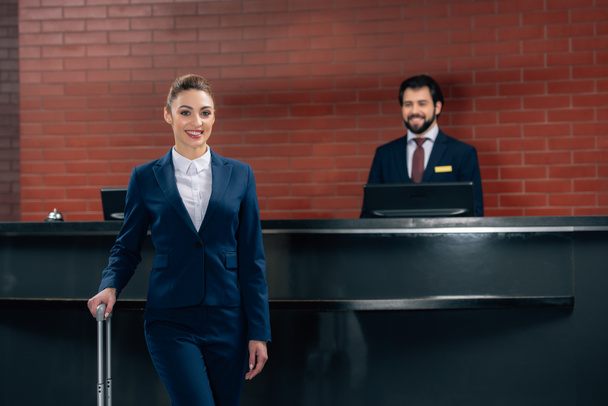 smiling businesswoman with luggage standing in front of hotel reception counter with administrator - Photo, Image