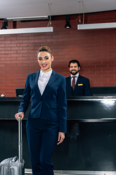 happy businesswoman with luggage standing in front of hotel reception counter with administrator - Fotoğraf, Görsel