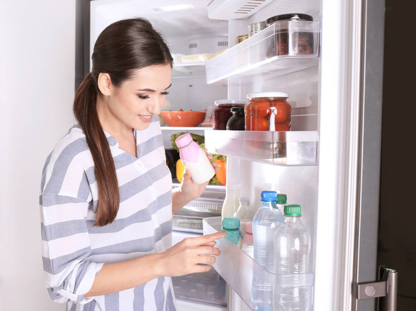 Young woman with bottle of yogurt near open refrigerator at home - Фото, зображення