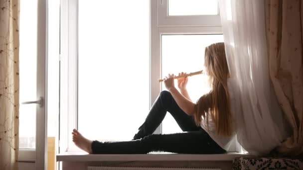 Young cute teen girl playing on flute sitting on windowsill at home - Footage, Video