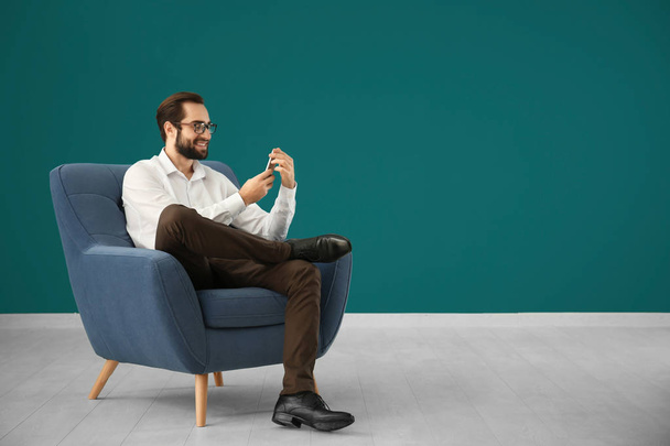 Handsome man with mobile phone sitting in comfortable armchair against color wall - Fotoğraf, Görsel