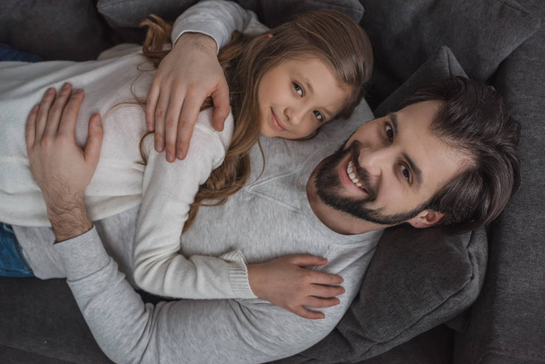 aus der Vogelperspektive: Vater und Tochter umarmen sich auf dem Sofa und blicken in die Kamera - Foto, Bild