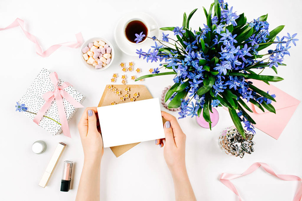 女性の手は青い春の花束、コーヒー カップ、お菓子、化粧品アクセサリーで白い背景に空のカードを保持します。 - 写真・画像