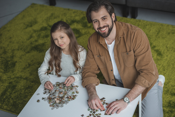 aus der Vogelperspektive: Vater und Tochter sitzen mit Puzzleteilen am Tisch und blicken in die Kamera - Foto, Bild
