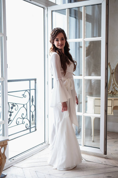 Beautiful bride in a white wedding peignoir in a morning. beauty portrait in interior - Foto, afbeelding