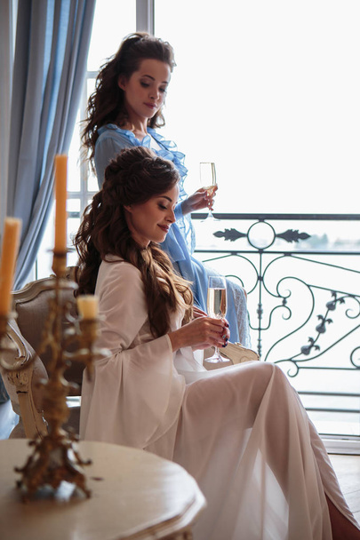 Beautiful twins young women with glasses of champagne in elegant interior room. bride's morning in a hotel - Foto, imagen