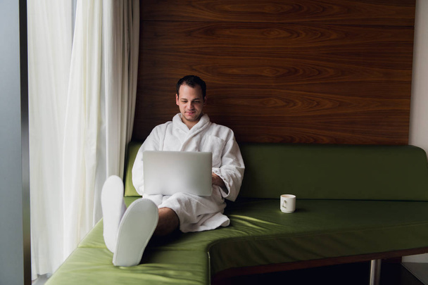 Joven hombre de negocios guapo con bata de baño en la cama con ordenador portátil en la habitación moderna. Persona autónoma que usa computadora portátil en el ático y mira la soleada vista de la ciudad en la ventana
 - Foto, imagen