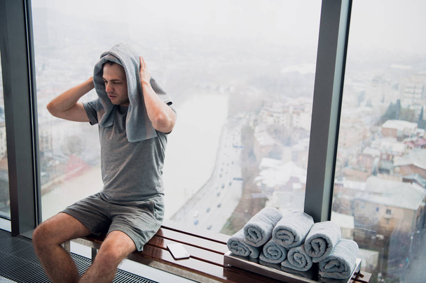 Un hombre joven y cansado sentado en un banco en un gimnasio. Sujetando teléfono móvil y toalla en su cabeza. Gimnasio en rascacielos con gran ventana y una gran vista
. - Foto, imagen