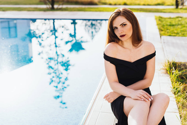 attractive beautiful woman in a black dress sits by the blue pool, enjoys and relaxes - Foto, Bild