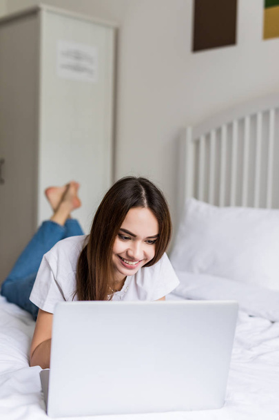 Mujer sonriente con las piernas levantadas levemente delante de su computadora portátil acostada en la cama
. - Foto, imagen