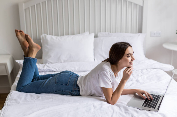 portrait of young american woman happy with laptop watching movie at white bed, rejoicing. concept of new technologies, advertising sales and freelance work - Photo, Image