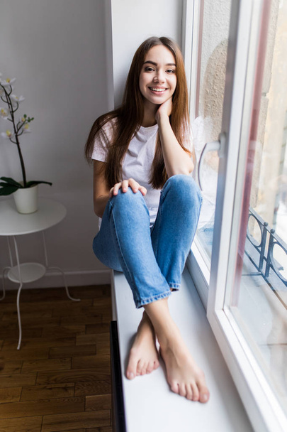 Ontspannend. Aantrekkelijke vrolijke jonge vrouw glimlachend en het dragen van een shirt en spijkerbroek en zittend op de vensterbank - Foto, afbeelding